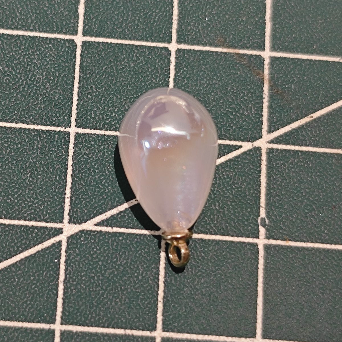 Close-up of a large white pearlescent mermaid teardrop charm with gold-plated loop on green cutting mat.