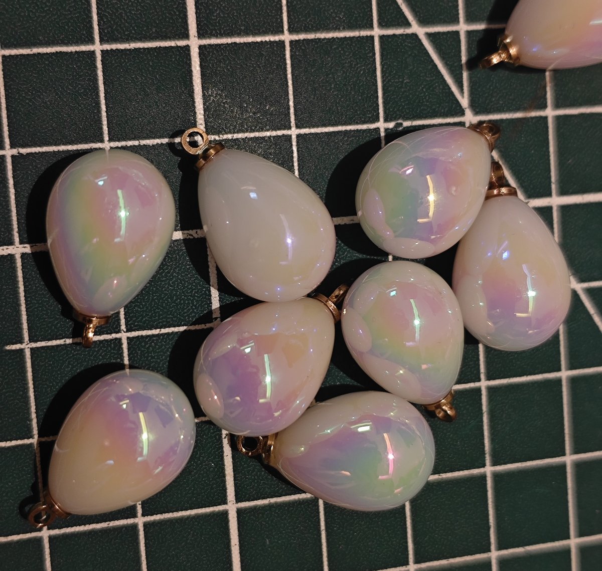 Close-up of multiple iridescent tear drop pendants with gold findings, showing rainbow color shift on green craft mat.