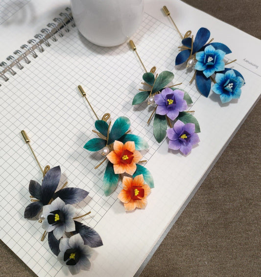 Ronghua handmade narcissus brooches in four colors — blue, purple, orange, and grey — each with velvet silk flowers, gold wire branches, and a pearl accent on a gold stick pin, displayed on white grid paper