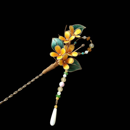 Ronghua handmade osmanthus hairpin with three golden yellow silk flowers, gold edge trim, dark green leaves, copper-wrapped stem, and long beaded chain drop ending in a white shell pendant, displayed on dark grey fabric