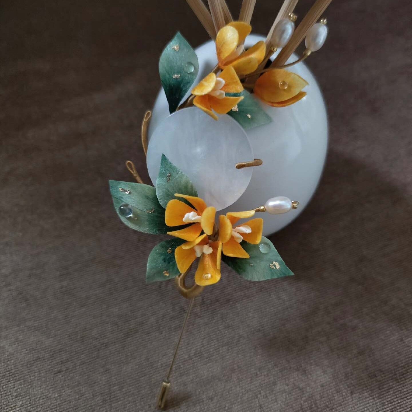 Ronghua handmade osmanthus brooch with golden yellow silk flowers, dark green leaves with gold foil, a large translucent jade disc, pearl sprigs, and a gold stick pin, displayed on white grid paper
