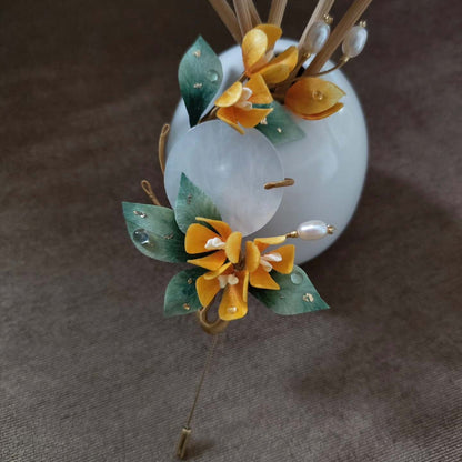 Ronghua handmade osmanthus brooch with golden yellow silk flowers, dark green leaves with gold foil, a large translucent jade disc, pearl sprigs, and a gold stick pin, displayed on white grid paper