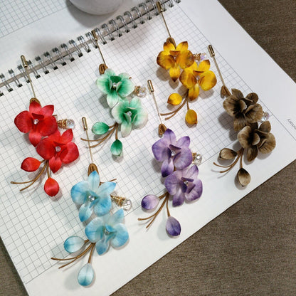 Ronghua handmade phalaenopsis brooches in six colors — red, blue, green, yellow-orange, purple, and taupe — each with crystal drop pendant and gold stick pin, displayed on white grid paper