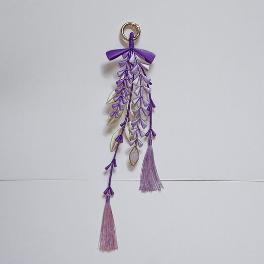 Handmade Chanhua wisteria pendant with cascading purple and lavender florets, cream-gold leaves, crystal leaf accent, purple bow, gold ring loop, and two pink silk tassels, displayed on a white background