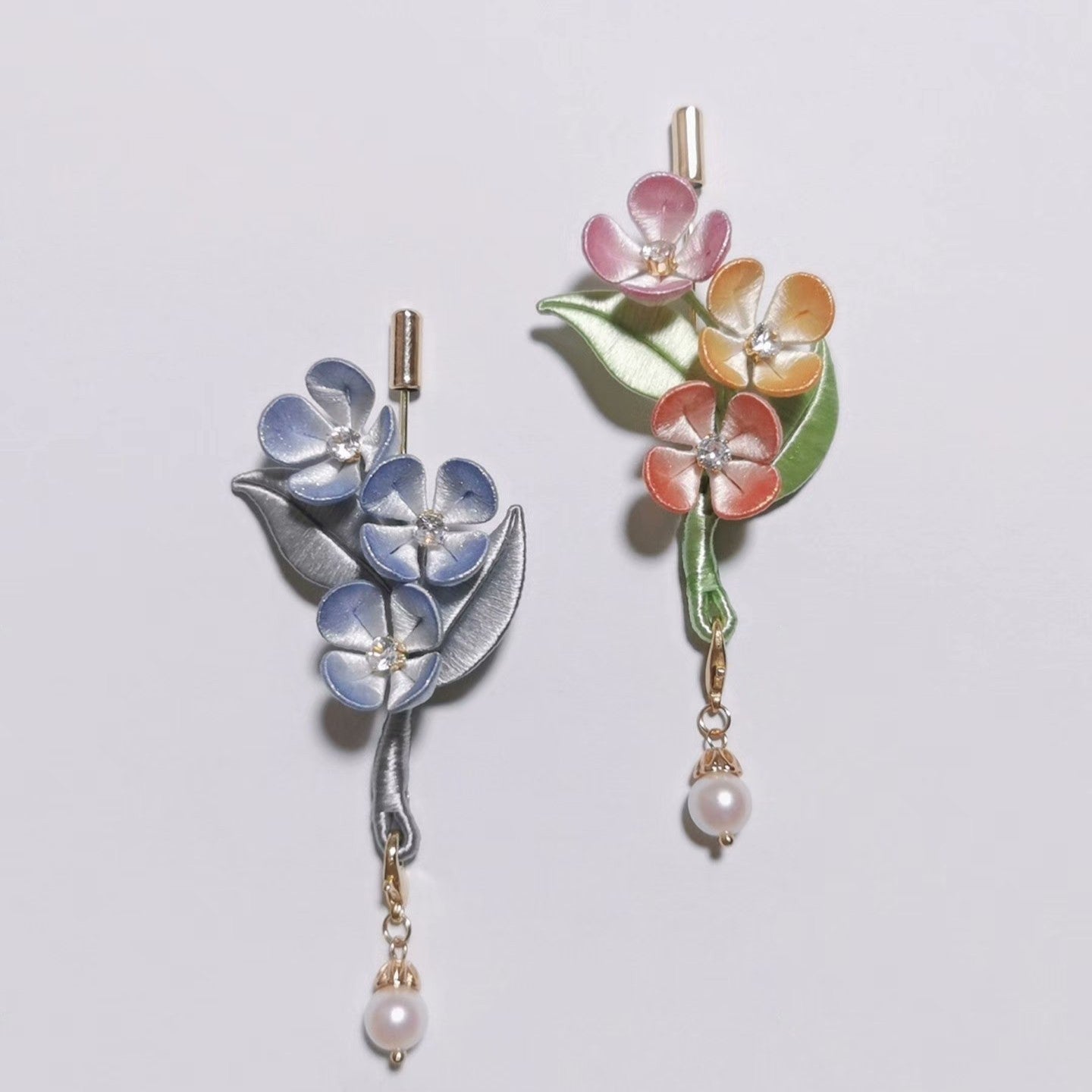 Chanhua Ruyan handmade flower brooches in two styles — blue with silver-grey leaves and pastel multicolor with green leaves — each with a pearl drop on a gold chain, displayed on a light background