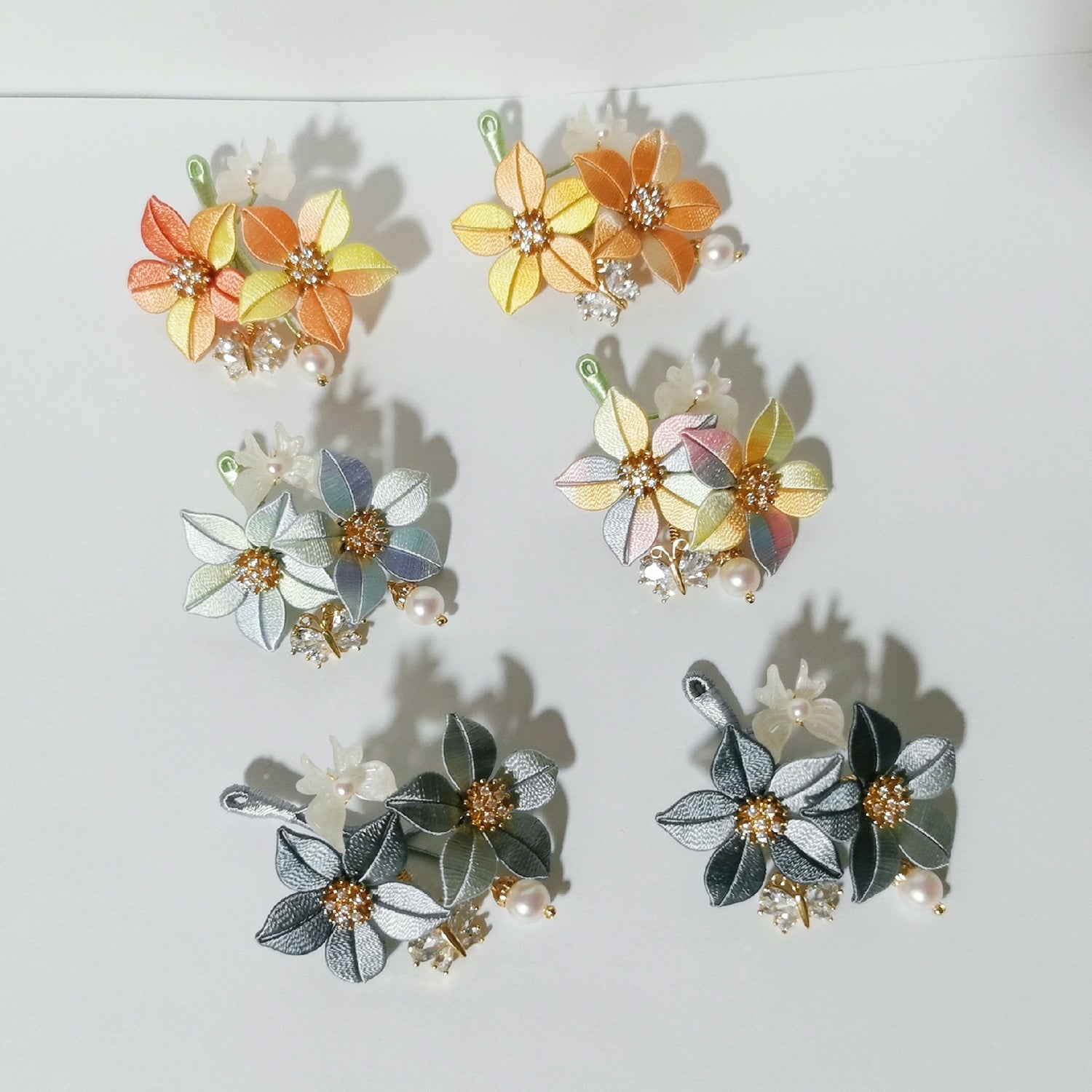 Chanhua Shijing handmade flower brooches in six color combinations including orange, yellow, blue-grey, pastel multicolor, dark grey, and charcoal, displayed on a white background