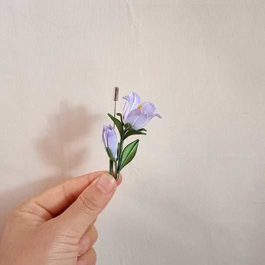 Chanhua handmade lavender bellflower brooch with open bloom, closed bud, dark green leaves, and fine stem on a stick pin, held in hand against a cream background