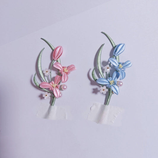 Chanhua handmade orchid brooches in pink and blue, each with green leaves, small white flower accents and pearl detail, displayed on a light purple background
