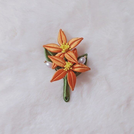Chanhua handmade orange flower brooch with two blooms, yellow stamens, green leaves, and pearl accents, displayed on white fabric