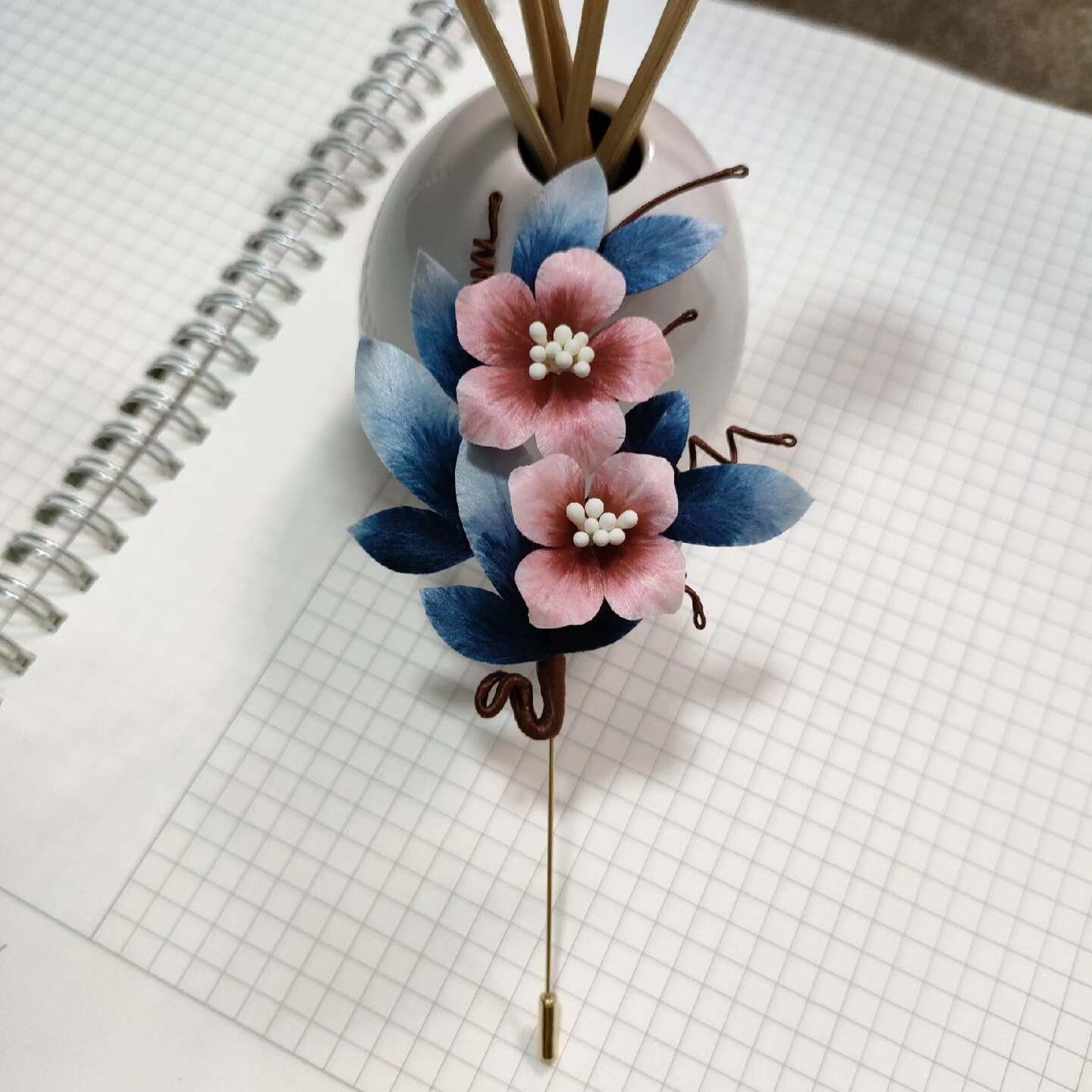 Ronghua handmade flower and leaf brooch with two pink silk blossoms, layered blue velvet leaves, dark wire curls, and a long gold stick pin, displayed on white grid paper