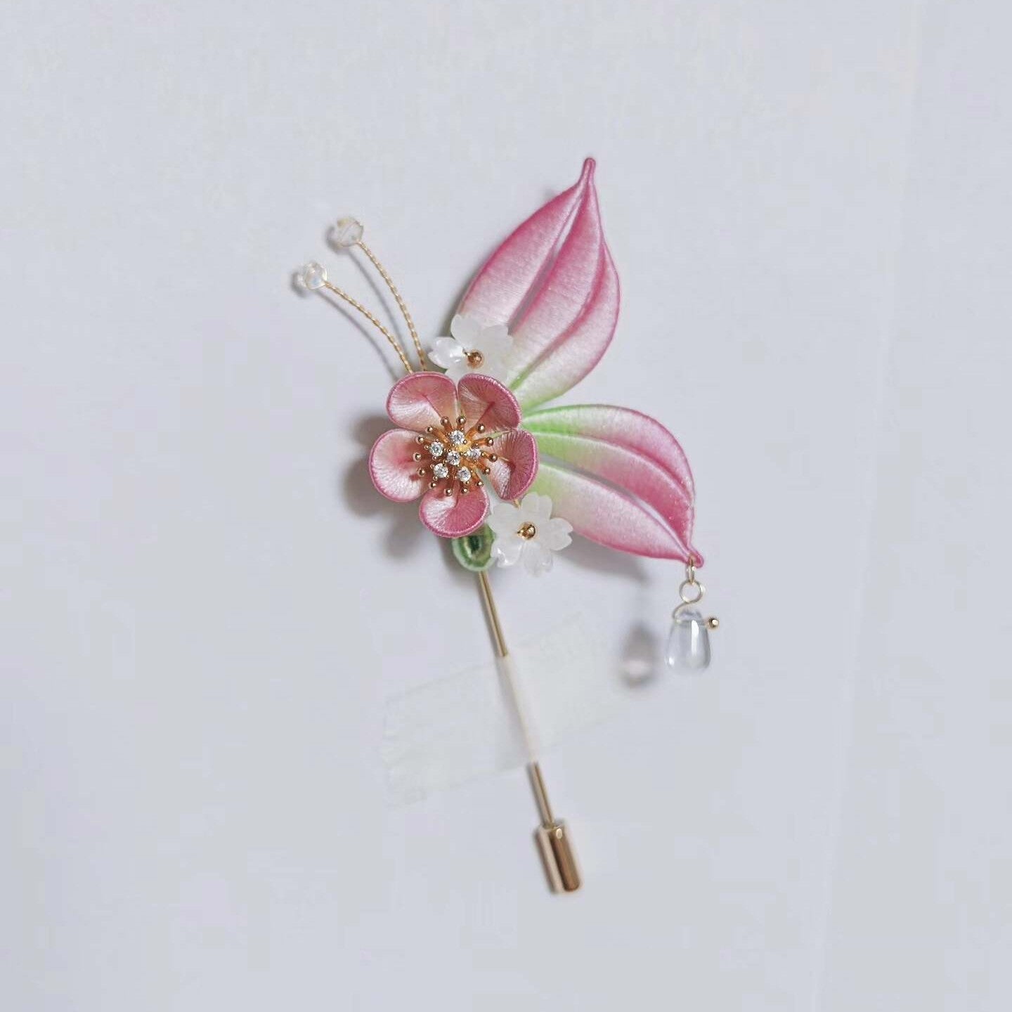 Chanhua handmade butterfly brooch with pink ombre wings, plum blossom body, gold antenna with crystal tips, and clear crystal drop pendant, displayed on a light gray background