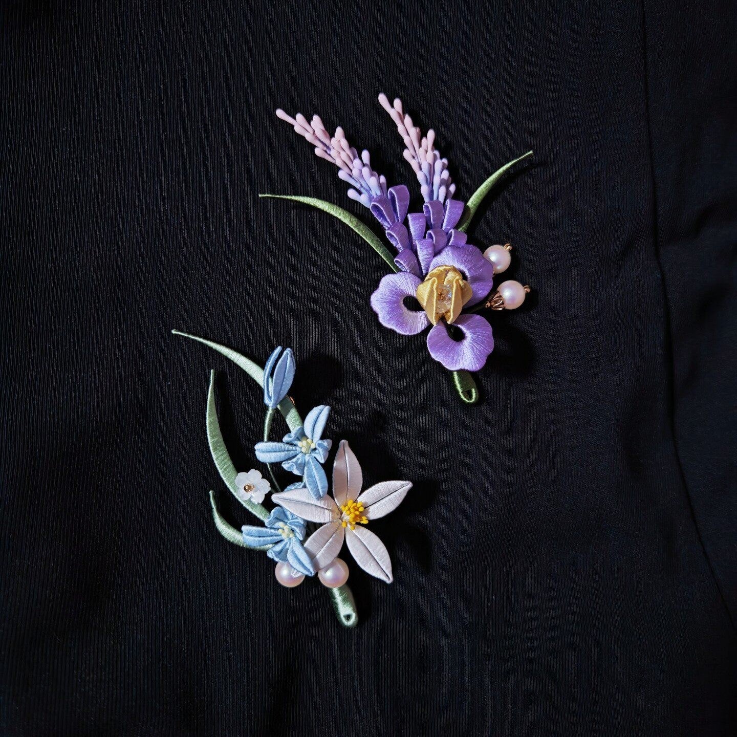 Chanhua handmade iris brooch and orchid brooch displayed side by side on a black jacket, showing purple iris with sage spikes and white orchid with blue buds