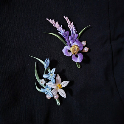 Chanhua handmade iris brooch and orchid brooch displayed side by side on a black jacket, showing purple iris with sage spikes and white orchid with blue buds