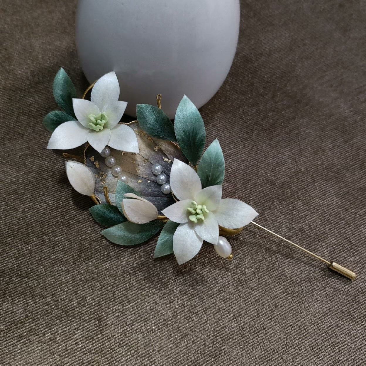 Ronghua handmade orchid brooch with two white silk blooms, green centers, teal leaves, a feather accent with gold foil, pearl clusters, and a long gold stick pin, displayed on dark grey fabric beside a white ceramic vase