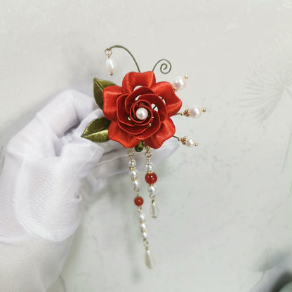 Chanhua Shiyi handmade red rose brooch with pearl center, gold-edge green leaves, and three long pearl and stone drop tassels, held in a white-gloved hand
