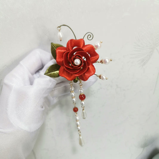 Chanhua Shiyi handmade red rose brooch with pearl center, gold-edge green leaves, and three long pearl and stone drop tassels, held in a white-gloved hand