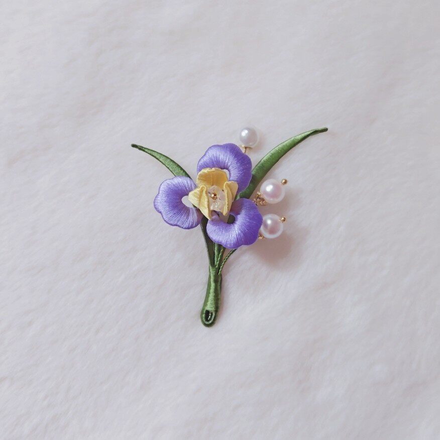 Chanhua handmade purple iris brooch with yellow center, green leaves, and three pearl accents on gold stems, displayed on white fabric