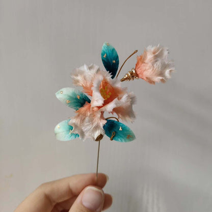 Ronghua handmade iris brooch with soft pink fringed petals, teal and turquoise velvet leaves with gold foil, yellow stamens, and a gold stick pin, held against a light grey background