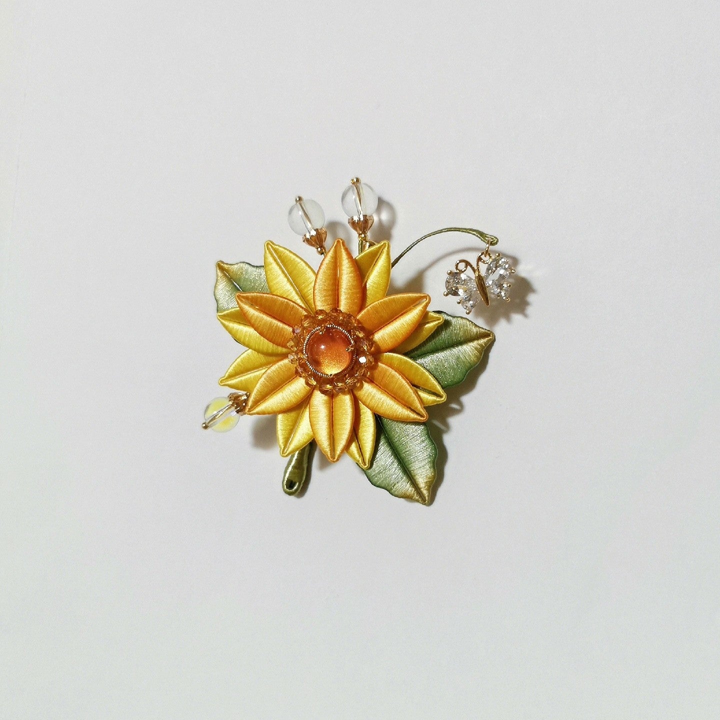 Chanhua handmade sunflower brooch with yellow petals, amber resin center, green leaves, moonstone bead accents, and crystal flower charm on a gold wire tendril, displayed on a light background