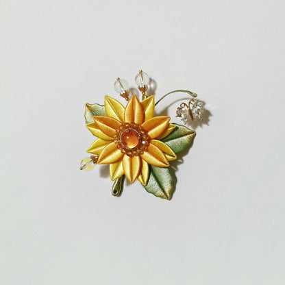 Chanhua handmade sunflower brooch with yellow petals, amber resin center, green leaves, moonstone bead accents, and crystal flower charm on a gold wire tendril, displayed on a light background