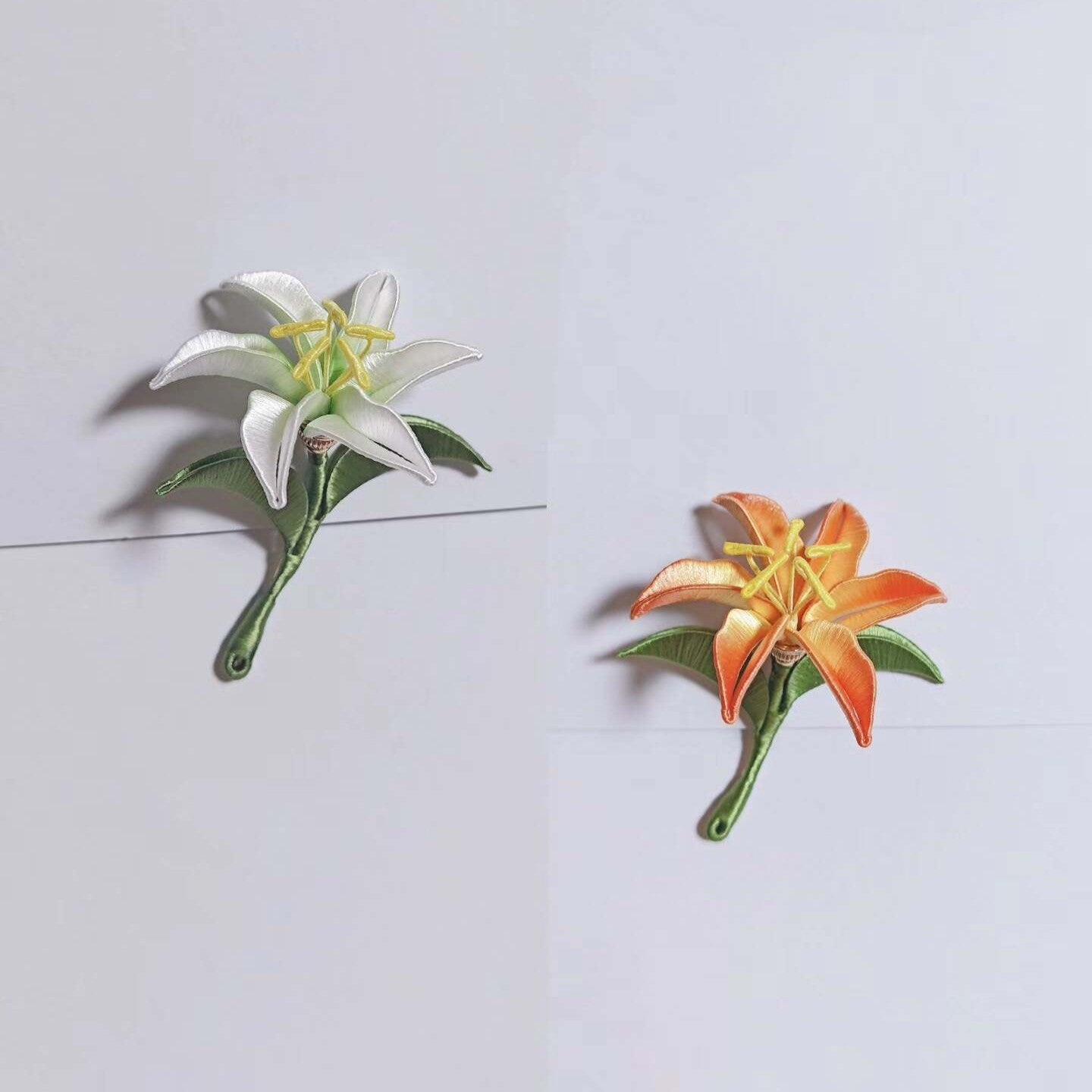 Chanhua handmade lily brooches in white and orange, each with yellow stamens and green leaves on a curved stem, displayed on a light gray background
