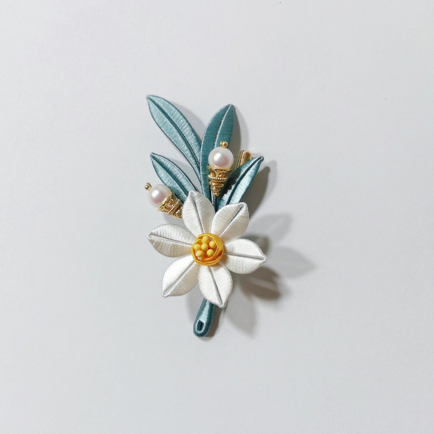 Chanhua handmade narcissus brooch with white petals, yellow center, teal-blue leaves, and two pearl accents on gold caps, displayed on a light gray background