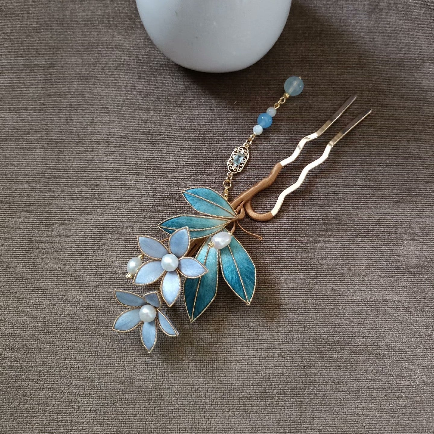 Ronghua Liyou handmade hairpin with teal velvet leaves, two soft blue flower blooms with pearl centers, gold filigree charm, light blue bead chain pendant, and gold U-fork pin, displayed on dark grey fabric