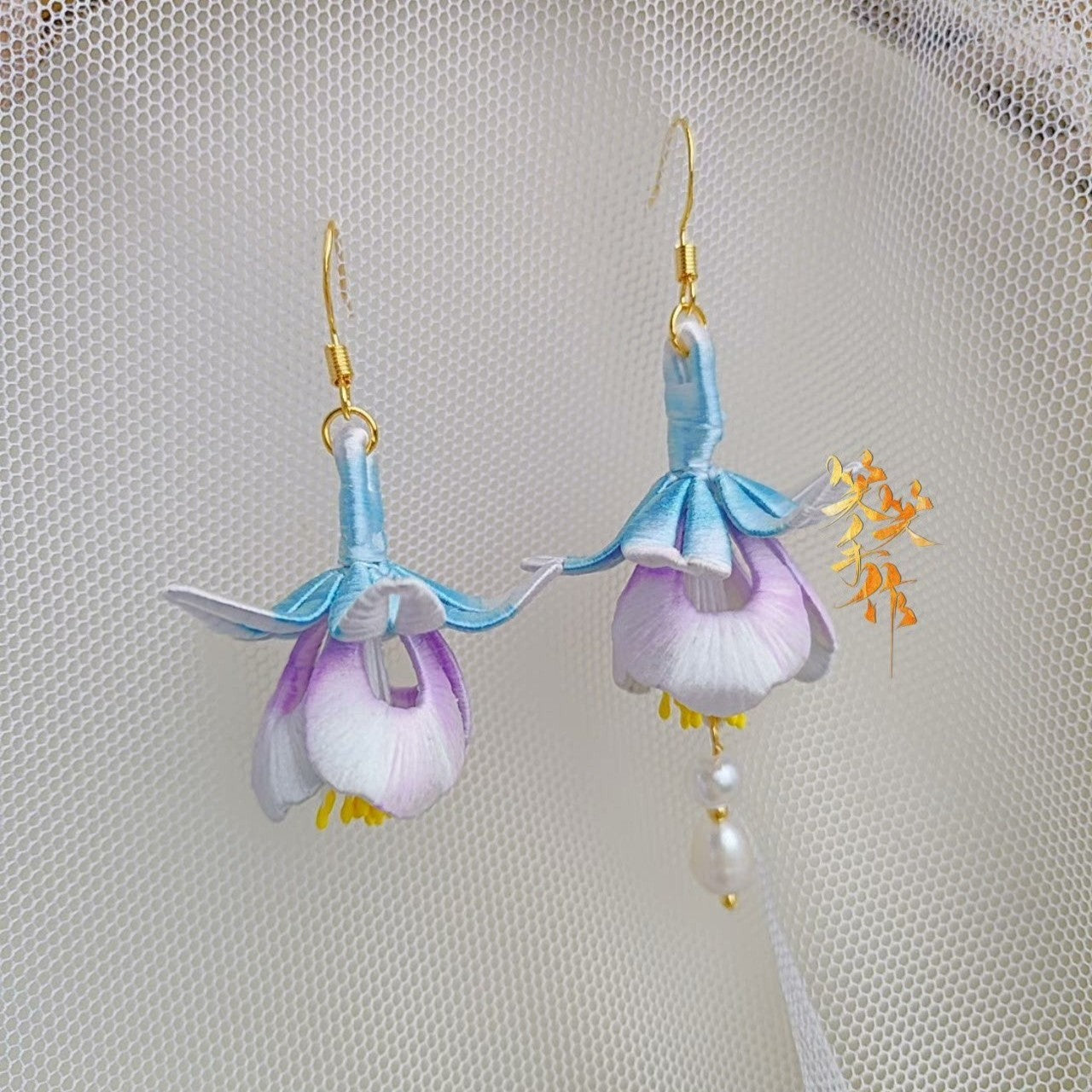 A pair of delicate, handcrafted Chanhua flower earrings with light blue and purple petals, showcasing the intricate details of the fabric and the 925 sterling silver ear hooks against a sheer white background.