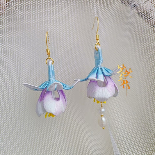 A pair of delicate, handcrafted Chanhua flower earrings with light blue and purple petals, showcasing the intricate details of the fabric and the 925 sterling silver ear hooks against a sheer white background.