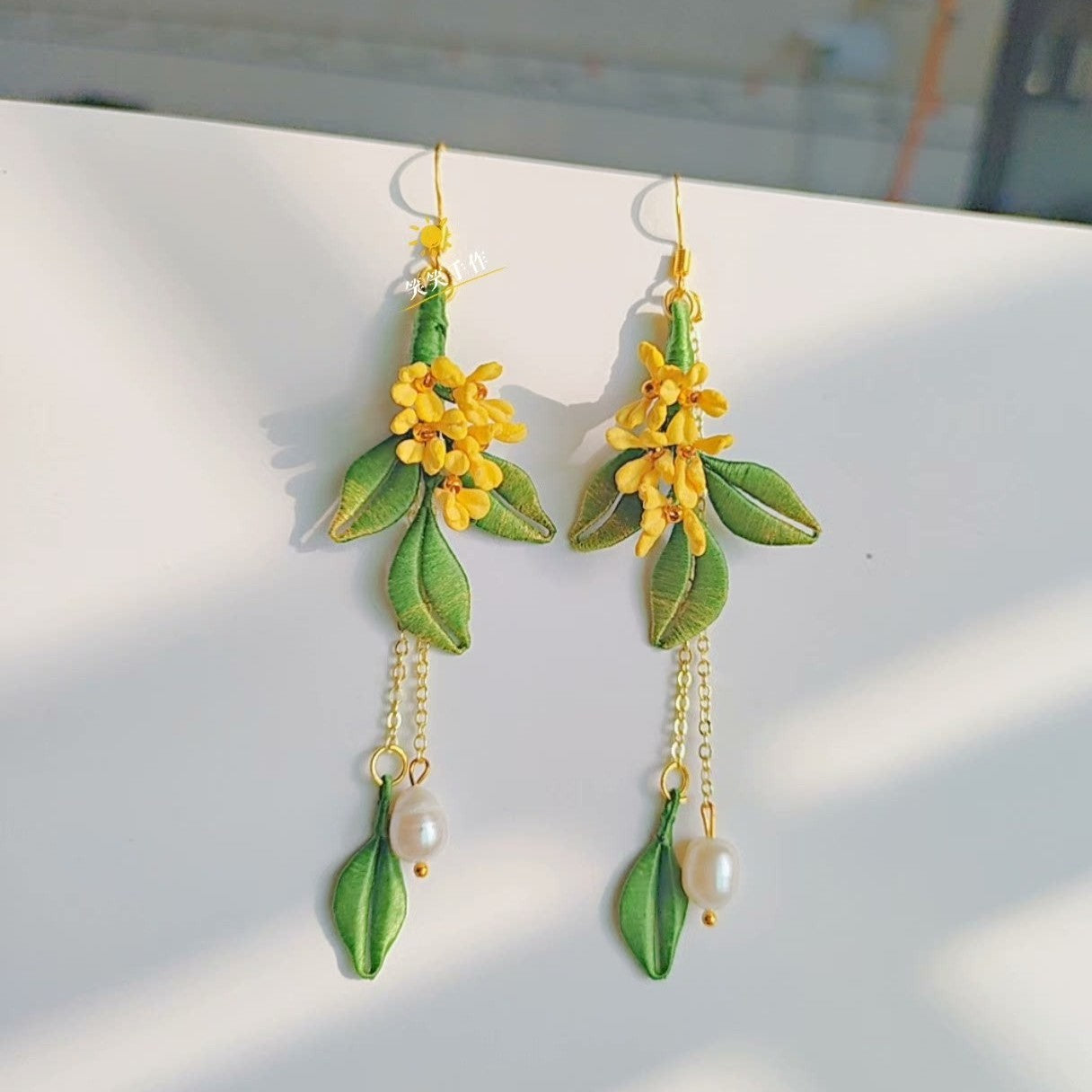 Handmade Chanhua osmanthus flower dangle earrings with yellow petals, green leaves, and white freshwater pearls on a white background.