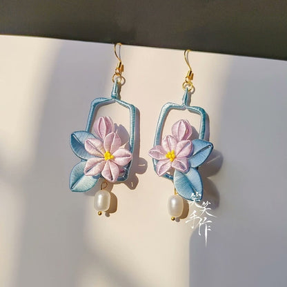 A pair of handcrafted Chanhua flower earrings with pink and purple petals, blue leaves, and natural freshwater pearl drops, hanging against a white background.