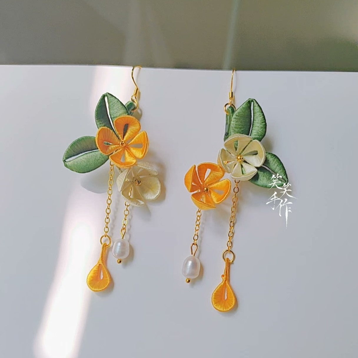 A pair of handmade Chanhua flower earrings featuring yellow and white fabric blossoms, green leaves, natural freshwater pearls, and 925 silver hooks, displayed against a white background.