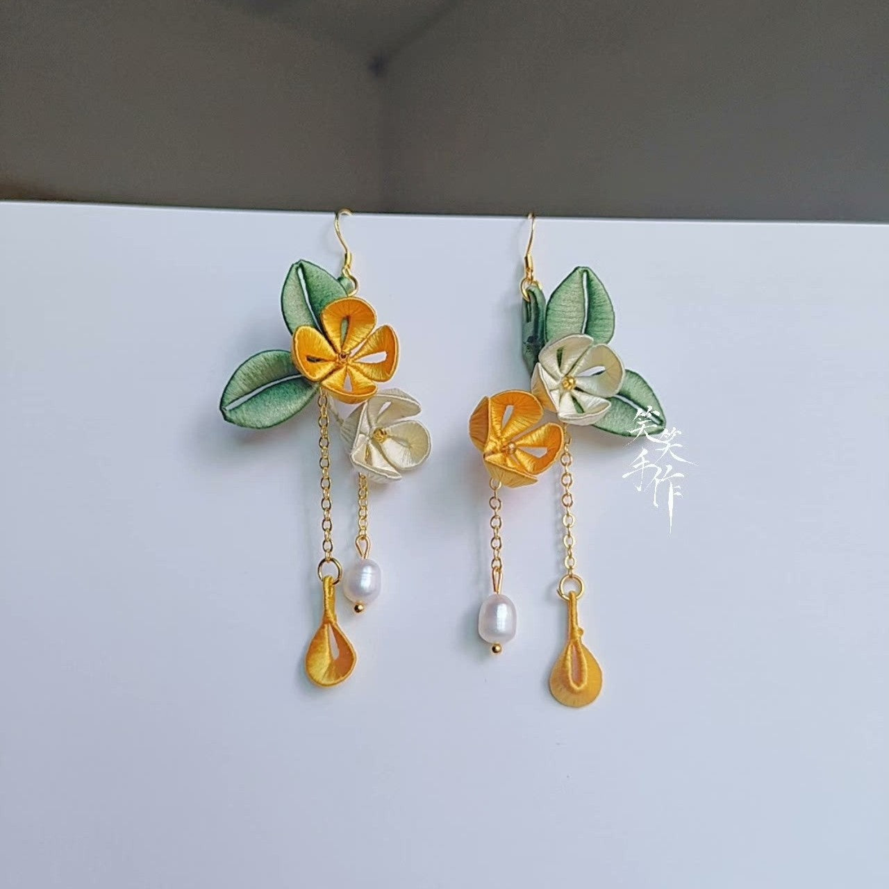Close-up of handmade Chanhua earrings showcasing intricate fabric details of yellow and white flowers with green leaves, dangling freshwater pearls, and golden tear-drop accents.
