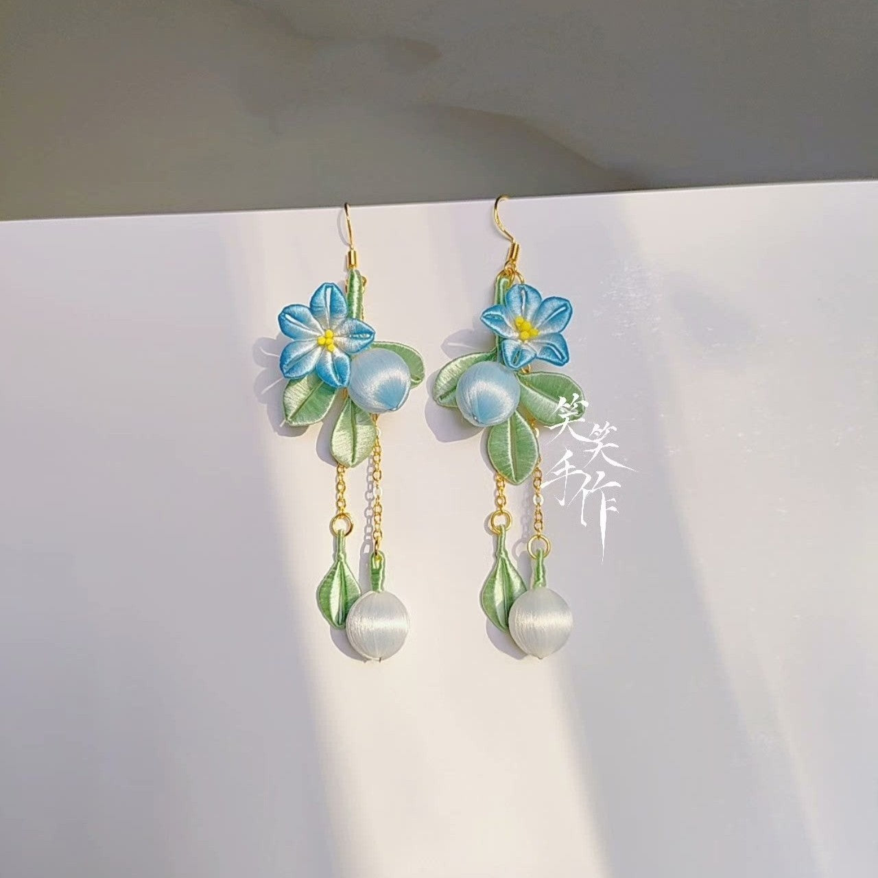 Close-up of intricate blue Chanhua fabric flowers, green leaves, and luminous white natural pearls on handmade dangle earrings.