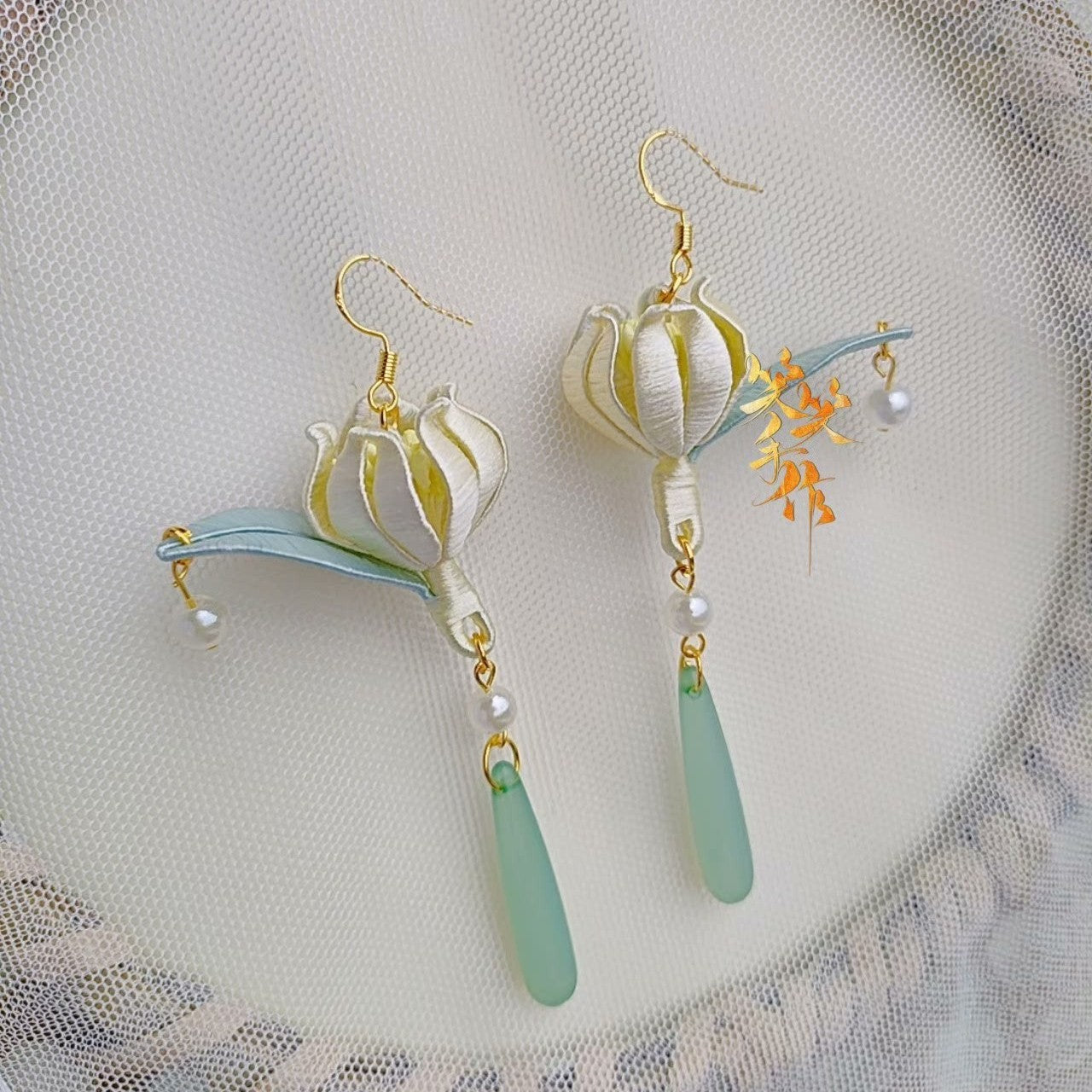 A pair of handcrafted white Chanhua bloom earrings with light blue leaves, natural freshwater pearls, and jade-like green drops, hanging on a sheer white fabric.