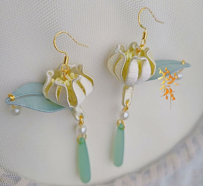 Front view of two handcrafted Chanhua bloom earrings, featuring delicate white and yellow folded fabric blooms, light blue leaves, lustrous pearls, and elongated green beads on a textured white background.