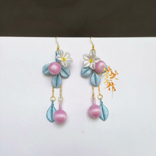 Close-up of a single handcrafted Chanhua earring showcasing intricate textile flowers, delicate blue leaves, and a pink dangling bead, with a golden ear hook visible.