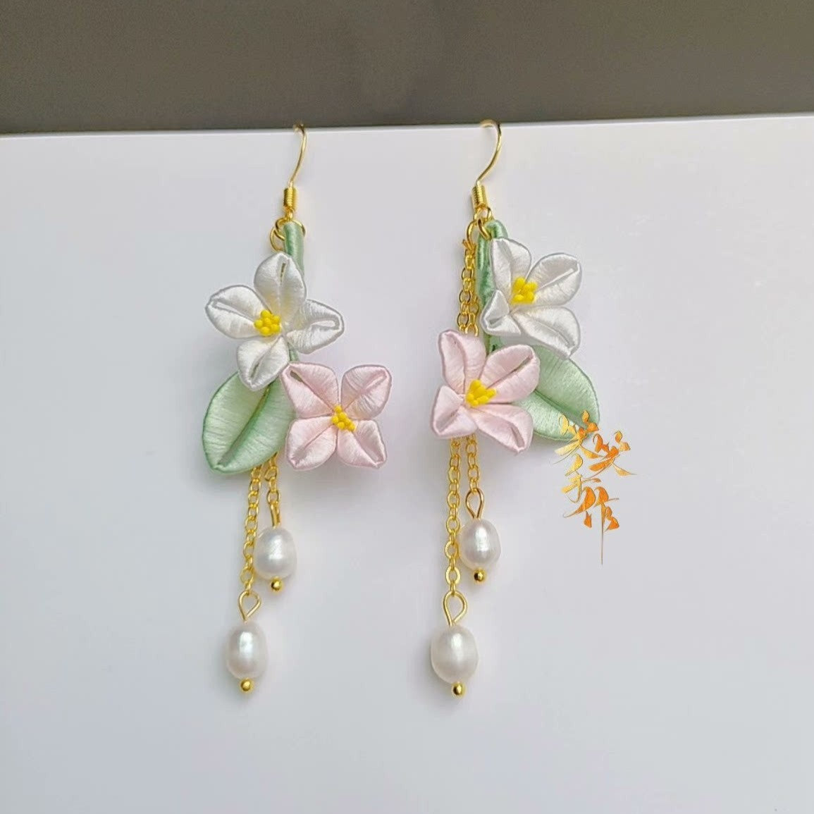 Handcrafted Chanhua flower earrings with white and pink blossoms, green leaves, and dangling natural freshwater pearls, hanging on a woven background.