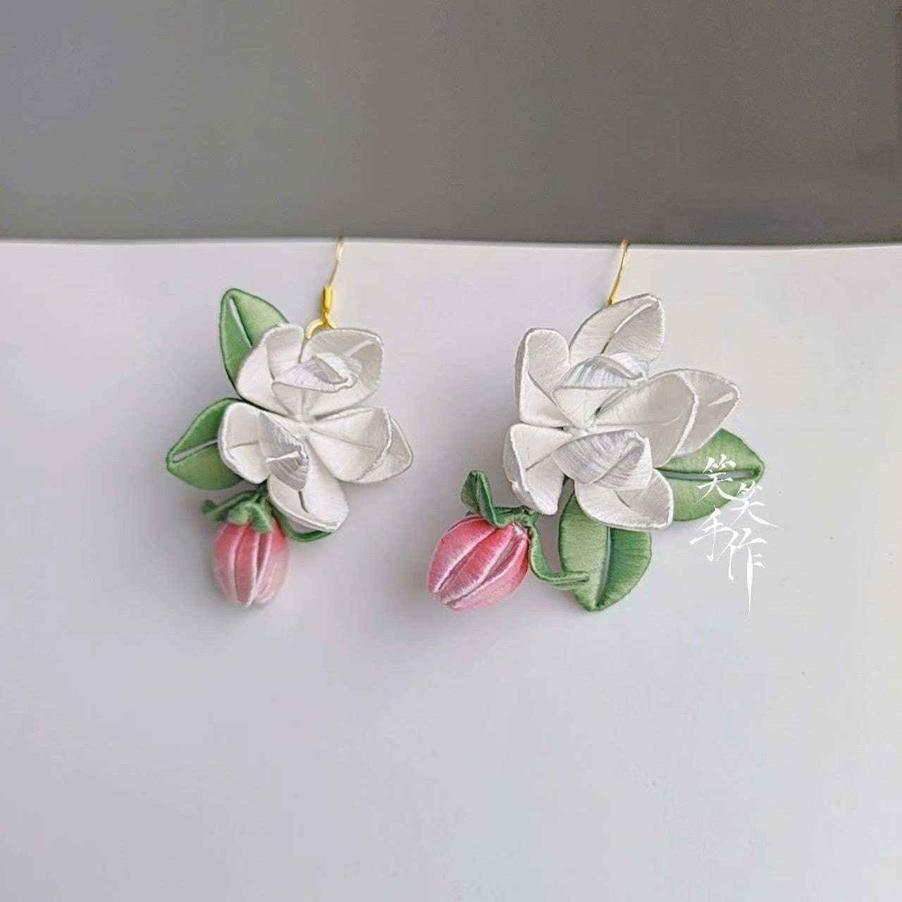 A pair of handcrafted white Chanhua flower and pink bud drop earrings with green leaves, made with delicate fabric and 925 sterling silver hooks, displayed against a light background.