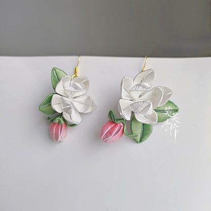 A pair of handcrafted white Chanhua flower and pink bud drop earrings with green leaves, made with delicate fabric and 925 sterling silver hooks, displayed against a light background.