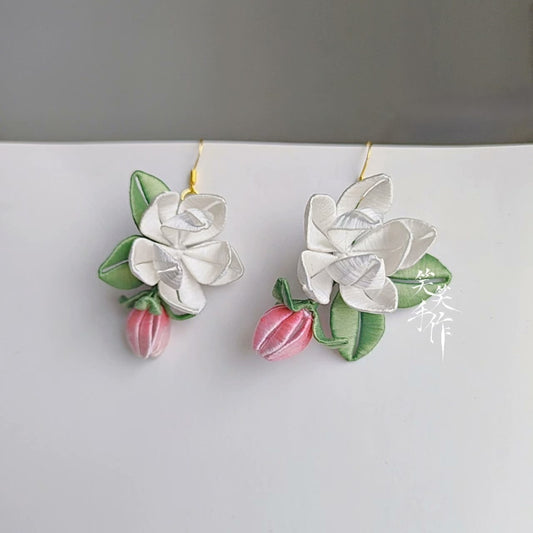 A pair of handcrafted white Chanhua flower and pink bud drop earrings with green leaves, made with delicate fabric and 925 sterling silver hooks, displayed against a light background.