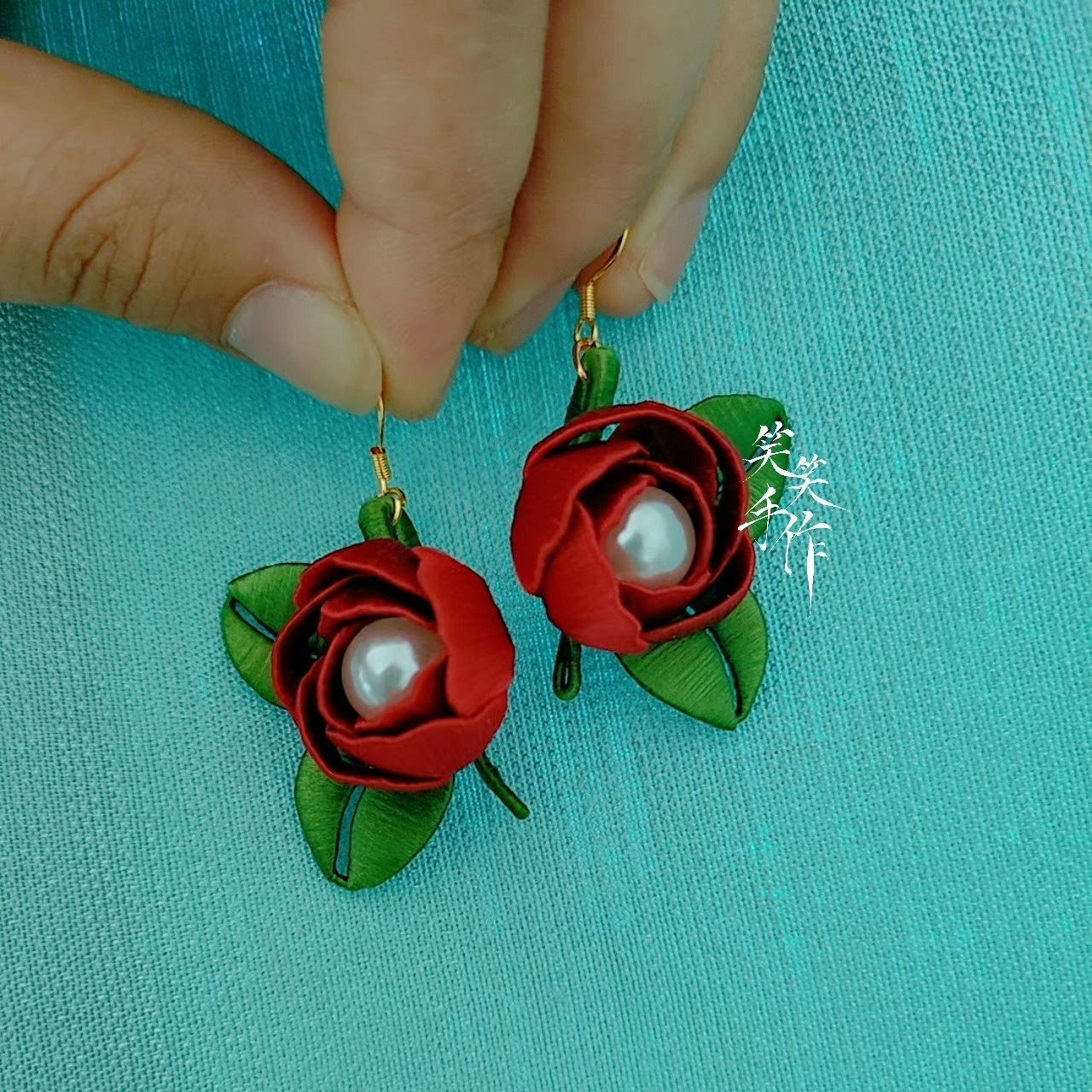A hand holding up a handcrafted Chanhua rose and pearl earring, showcasing its size and detailed craftsmanship against a blue background.