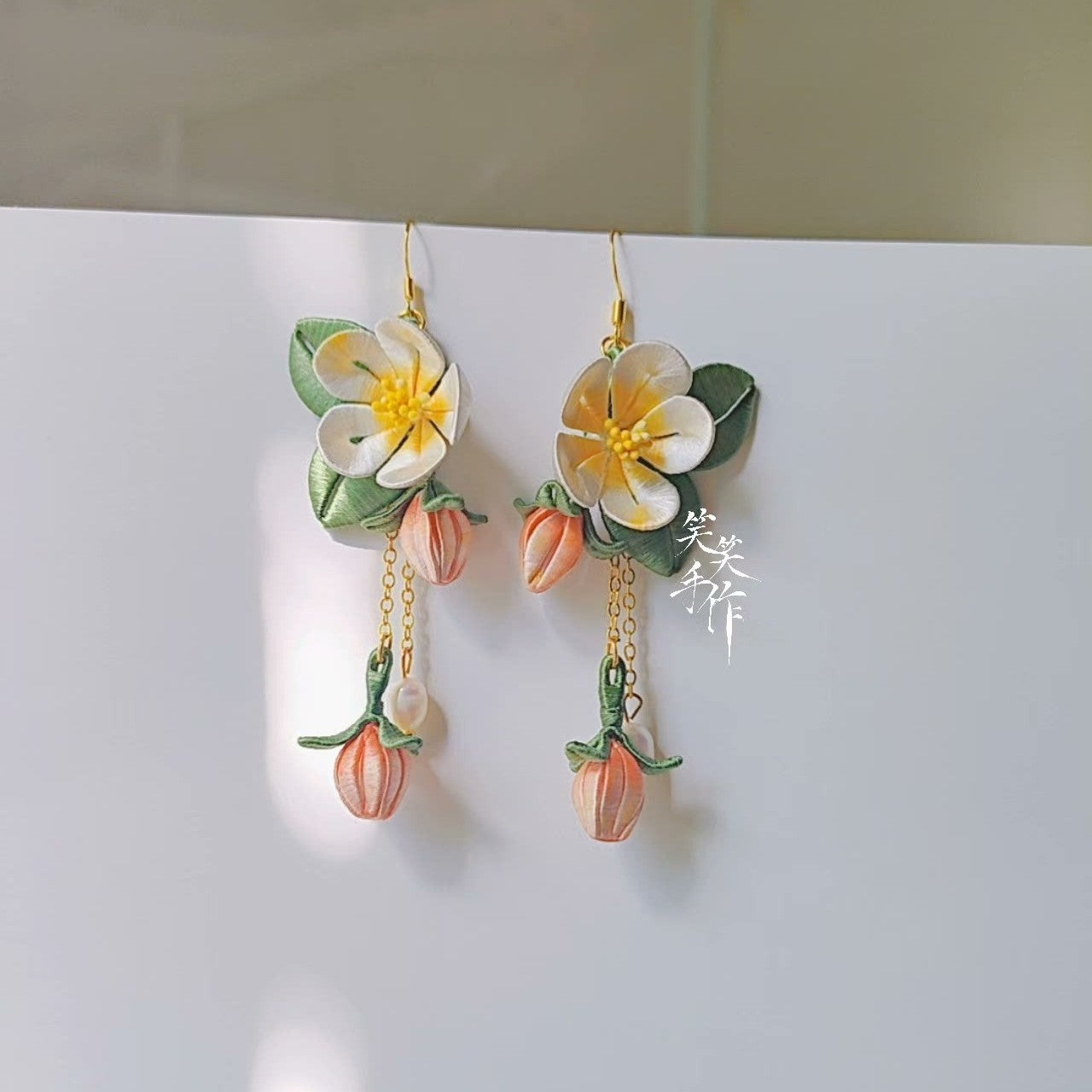 Close-up of handcrafted white and yellow camellia flower earrings with green leaves, dangling peach-colored buds, and natural freshwater pearls, showcasing intricate detail against a white background.