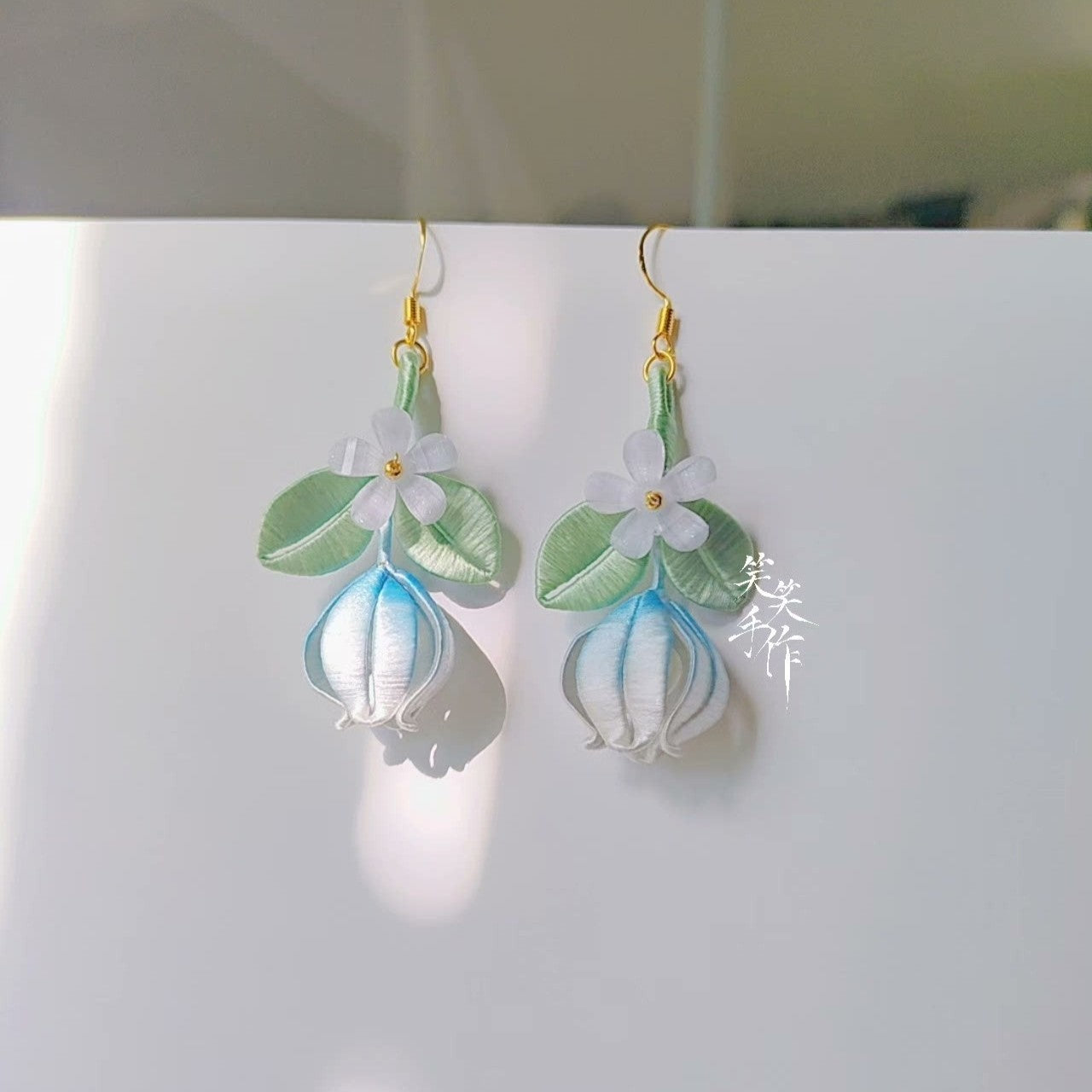Close-up shot of a pair of handcrafted Chanhua earrings, highlighting the intricate thread-wrapping details of the white flowers, green leaves, and blue-gradient buds, with sunlight casting shadows.