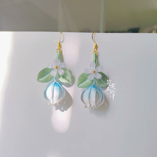 Close-up shot of a pair of handcrafted Chanhua earrings, highlighting the intricate thread-wrapping details of the white flowers, green leaves, and blue-gradient buds, with sunlight casting shadows.