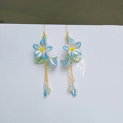 Close-up of a pair of handmade Chanhua flower dangle earrings with blue and white petals, yellow centers, and small green beads on 925 silver hooks, displayed against a white background.