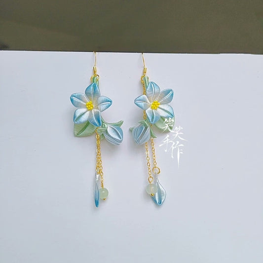 Close-up of a pair of handmade Chanhua flower dangle earrings with blue and white petals, yellow centers, and small green beads on 925 silver hooks, displayed against a white background.