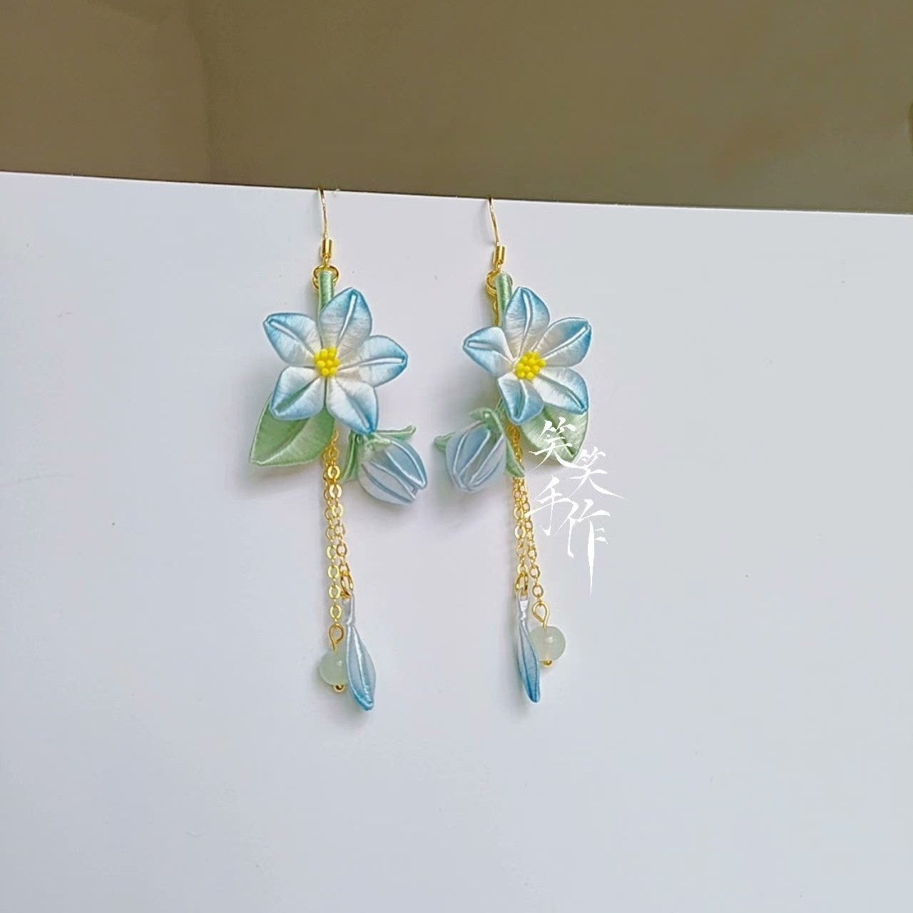 Full view of elegant handmade Chanhua flower earrings featuring intricate silk artistry, blue and green accents, and gold-tone chains with small jade-like beads, suspended for display.