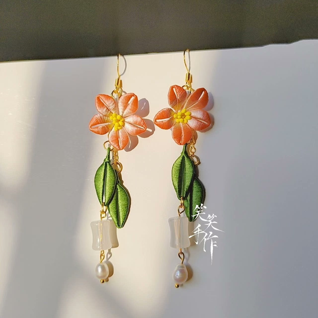 Close-up of handcrafted Chanhua flower and green leaf drop earrings with natural freshwater pearls and white beads, showcasing intricate detail and vibrant orange and green colors.