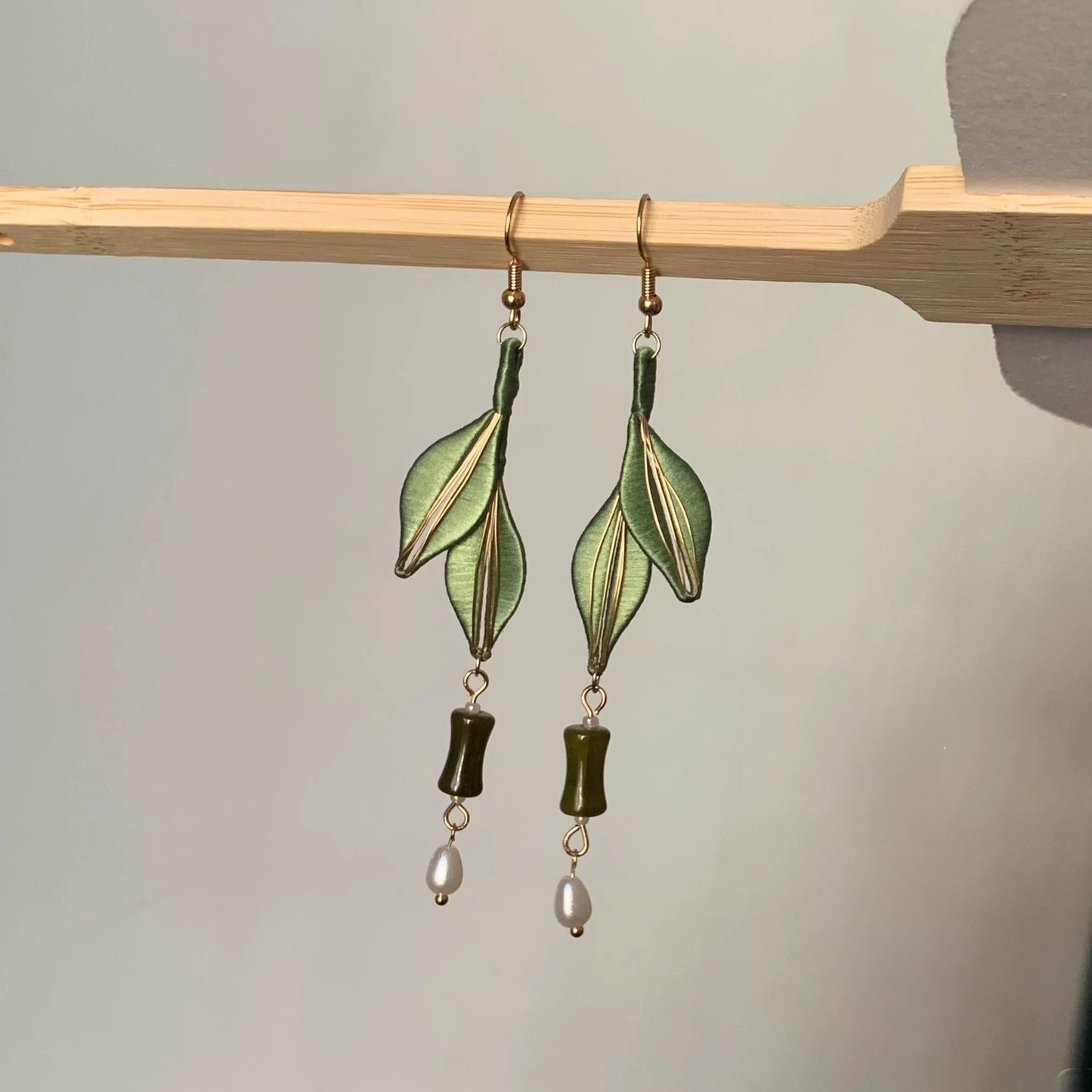 A close-up shot of the handcrafted Chanhua earrings with dark green leaves, a jadeite bead, and a pearl, hanging on a wooden stick.